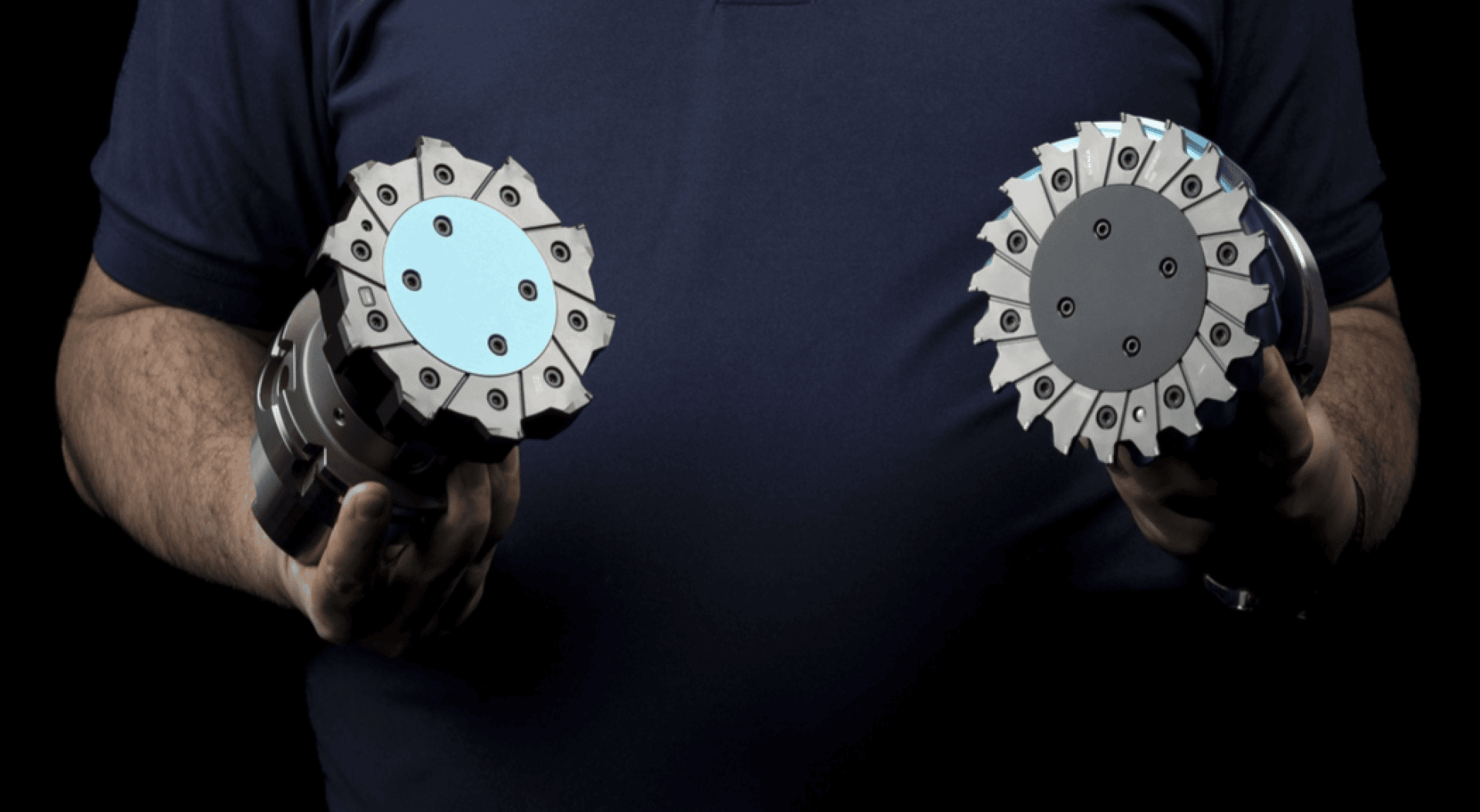 Eine Person in einem dunkelblauen Hemd hält vor einem schwarzen Hintergrund zwei industrielle Schneide- oder Fräswerkzeuge mit mehreren Metallzähnen, eines in jeder Hand.
