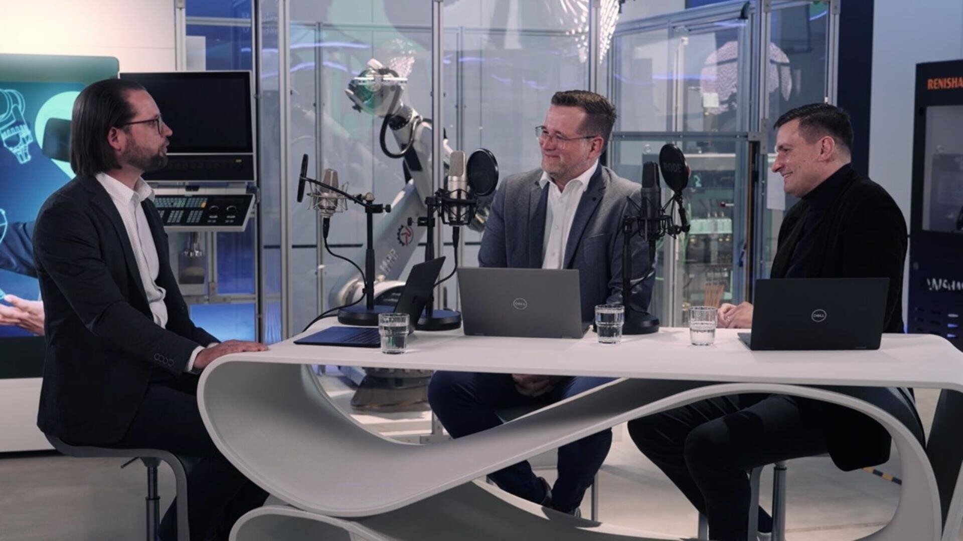 Drei Männer in Anzügen sitzen an einem geschwungenen weißen Tisch mit Mikrofonen und Laptops und führen eine Diskussion. Im modernen, gläsernen Hintergrund sind ein Roboterarm und elektronische Geräte zu sehen.