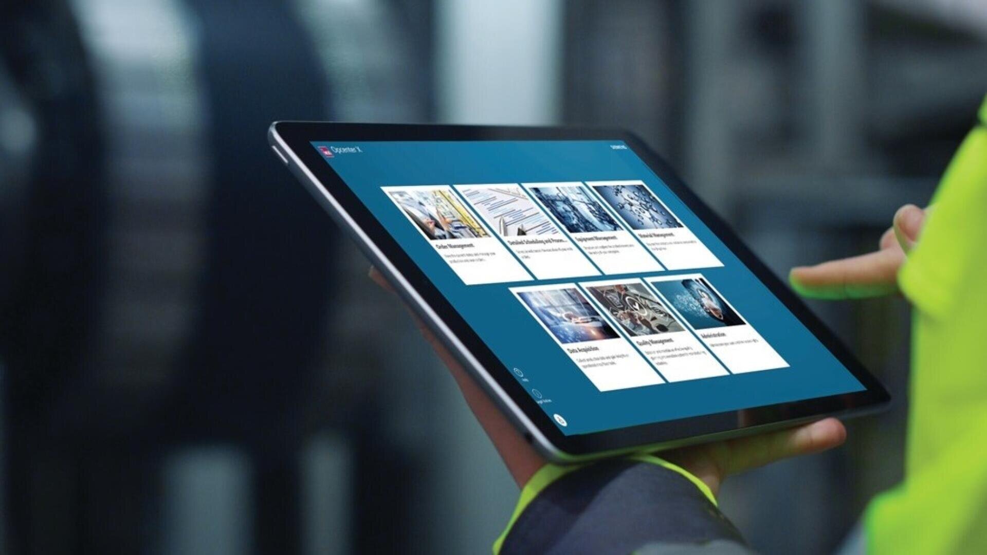 Eine Person in einer gelben Jacke hält und benutzt ein Tablet, das eine blaue Oberfläche mit verschiedenen Bildern und Textfeldern anzeigt, die möglicherweise mit industriellen oder technischen Themen zu tun haben. Im Hintergrund sind unscharfe Maschinen zu sehen.