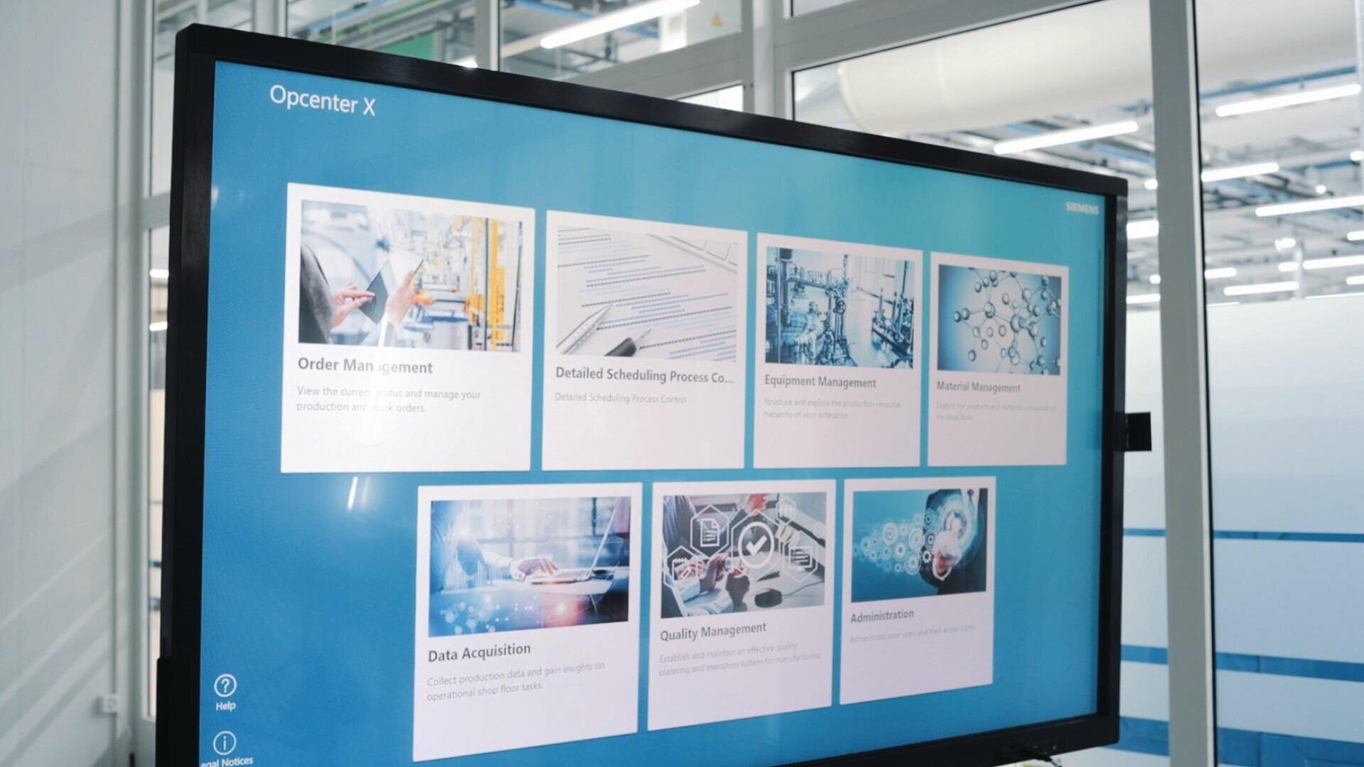 Ein großer Monitor zeigt die Softwareoberfläche von Siemens Opcenter X mit sechs Modulen, darunter Order Management, Equipment Management und Quality Management, in einer modernen Industrieumgebung.